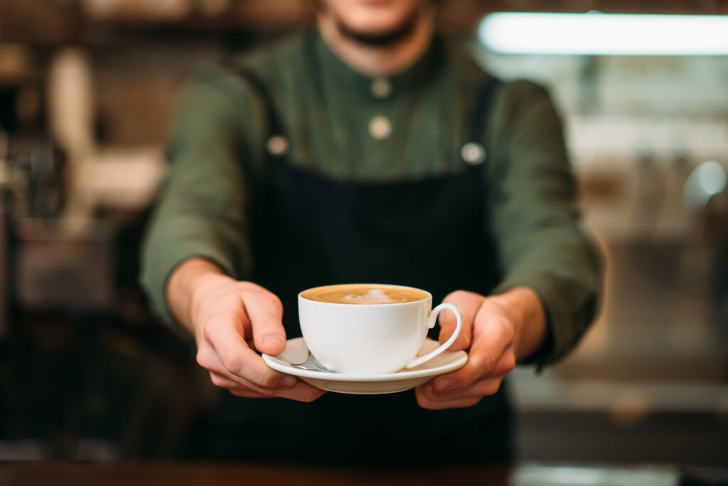 Waiter in black apron stretches a cup of coffee with cream in hands.